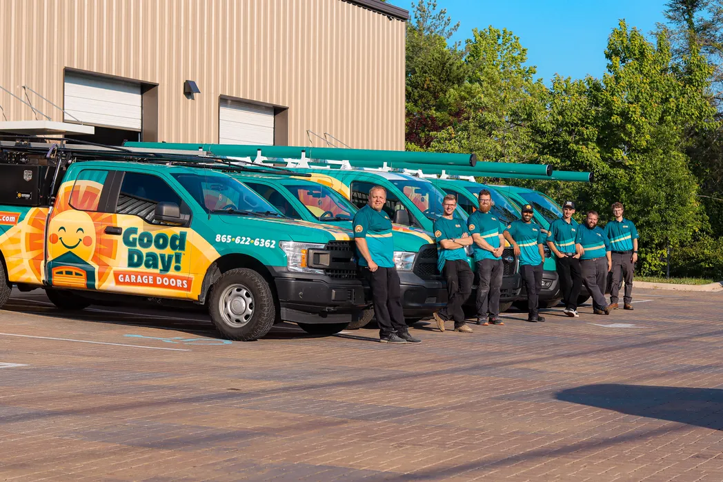 Seven Good Day Garage Doors technicians in uniform in front of a fleet of wrapped service pickup trucks.